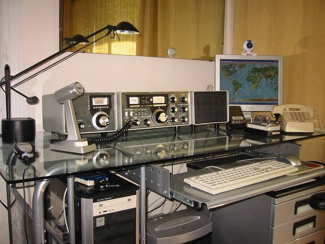 Masa's Yaesu station transceiver, external VFO, matching speaker, microphone all sitting on a modern glass desktop with keyboard drawer for his computer keyboard.